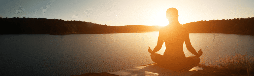 Rolig silhuet af en person der mediterer ved en sø i solnedgang, som symboliserer indre ro, nærvær og mental balance.